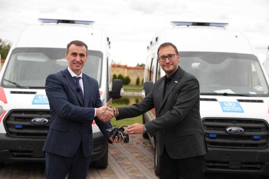 Two men in suits shaking hands in front of two white ambulances, with a building and trees visible in the background.