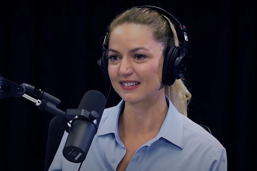 A woman wearing a light blue button-up shirt and black headphones, speaking into a microphone in a recording studio setting.