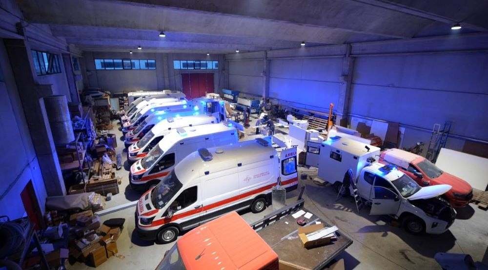 A garage or warehouse filled with multiple white and red emergency response vehicles, likely ambulances, parked in a row.