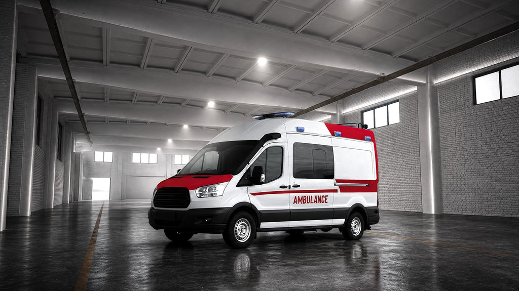 An ambulance parked in a large, empty warehouse or garage with a high ceiling and exposed beams, featuring a white body with red stripes and the word "AMBULANCE" written on the side in red letters.