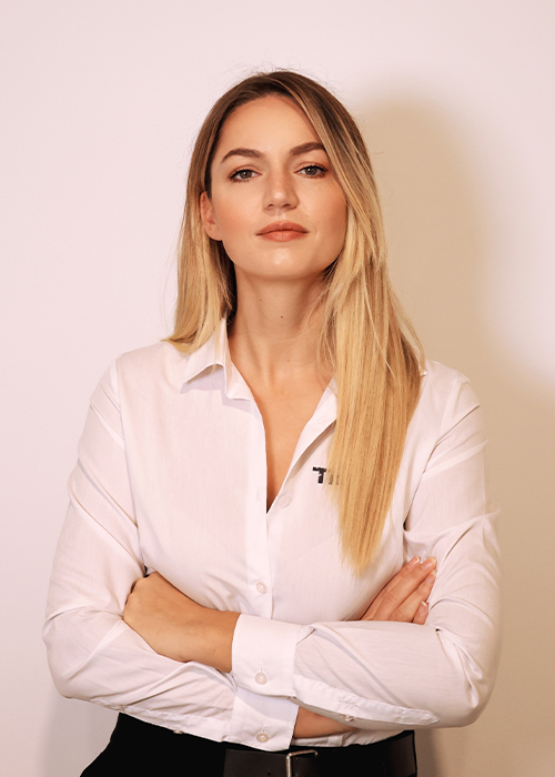 A woman posing in a white button-down shirt and black pants, with her arms crossed in front of a plain white wall.