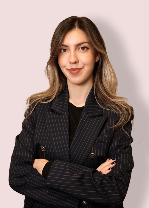 Here is a concise description of the image in one sentence: The image depicts a young woman with long brown and blonde hair, wearing a black pinstripe suit jacket, standing in front of a light pink background.