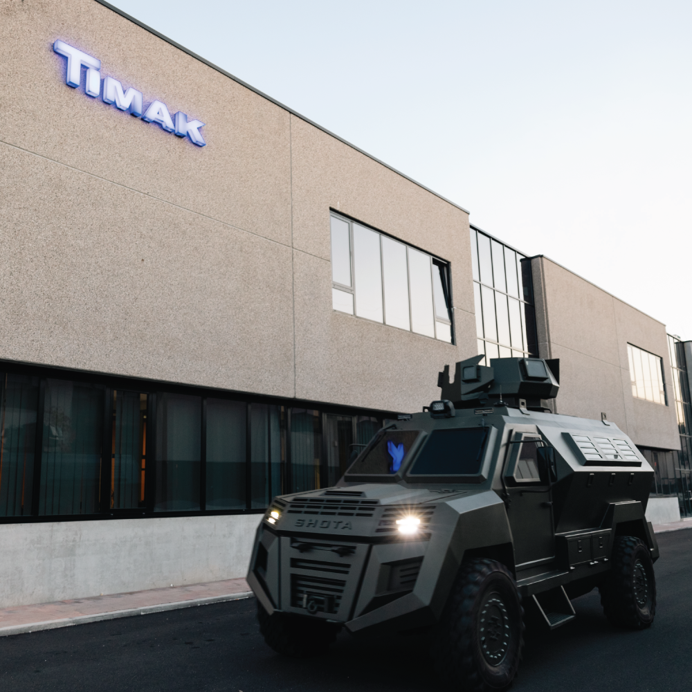 A military-style vehicle, likely an armored personnel carrier, parked in front of a building with the word "Timaik" displayed on it, set against a gray background.