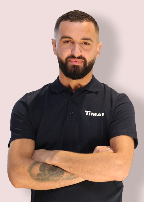 A man in a black polo shirt with the word "TIMAI" on the left side of his chest, standing against a gray background.