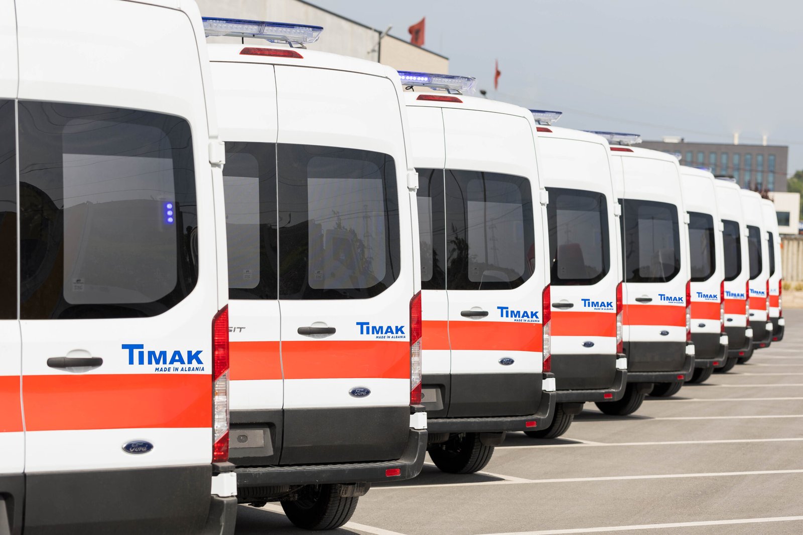 A row of white vans with orange and blue stripes, parked in a lot with a building in the background, suggesting a commercial or industrial setting.