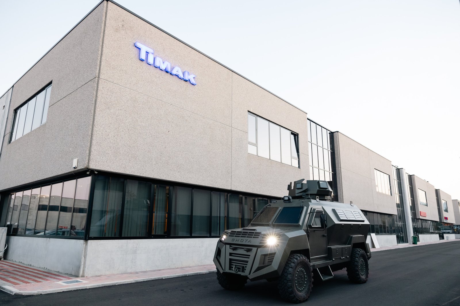 A silver, rugged, off-road vehicle, likely an armored personnel carrier or military vehicle, situated in front of a large, modern building with the word "TIMAC" displayed prominently on its side.
