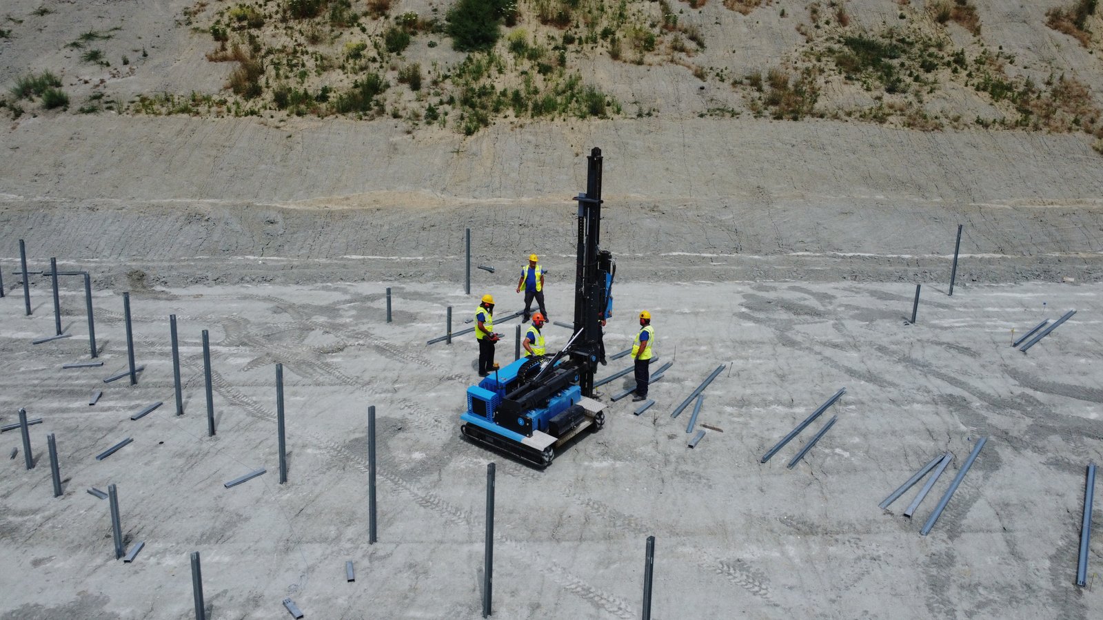 A blue and black drilling rig on a construction site, surrounded by metal rods in the ground.