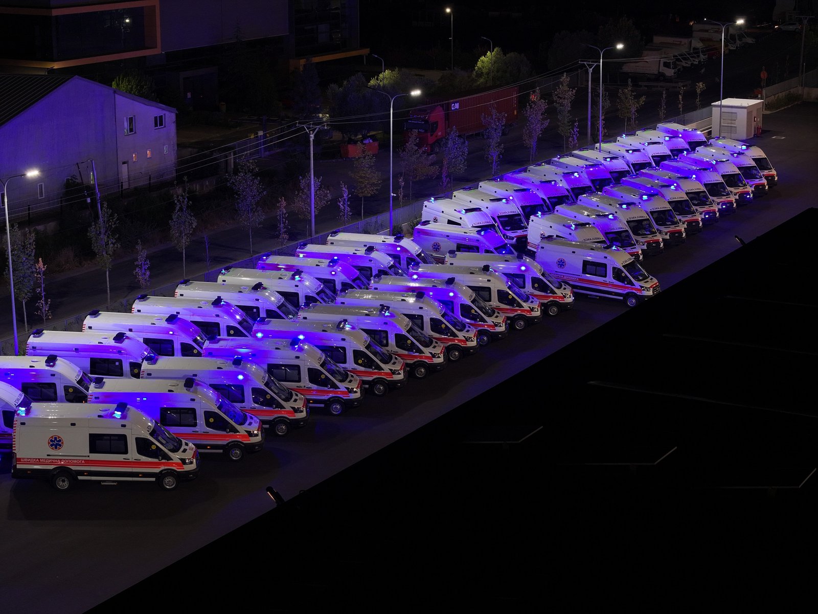 A nighttime scene of a long line of white and red ambulances parked on the side of a road.