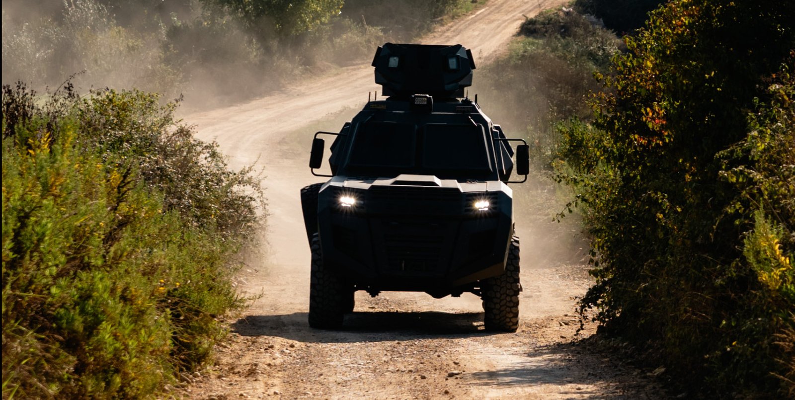 A dark-colored, four-wheel-drive military vehicle driving down a dusty dirt road surrounded by green foliage.