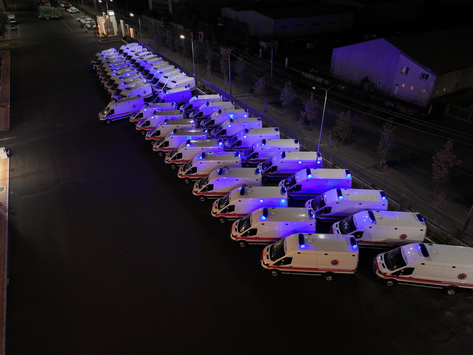 A line of white ambulances with blue lights parked in a lot at night, with a building in the background.