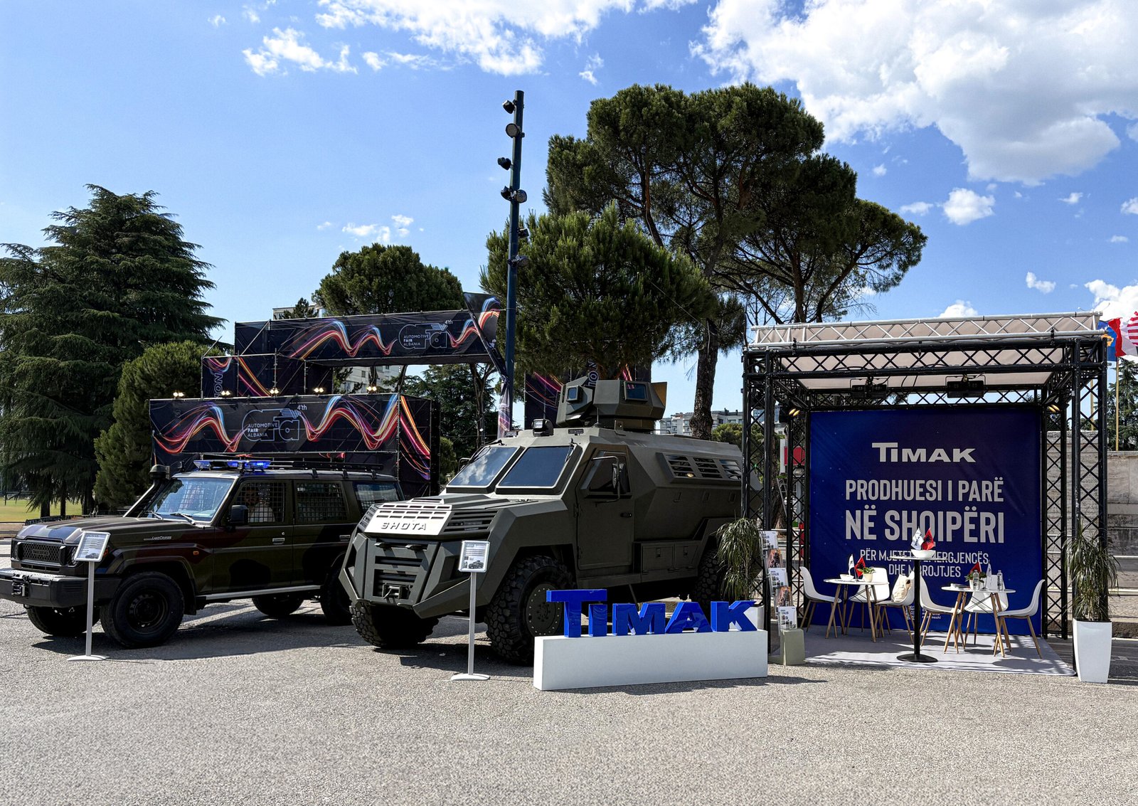 A military vehicle parked in front of a blue banner with white text, situated on a gray surface. The vehicle is dark green and features a large windshield and a turret on top. It is positioned in front of a blue banner with white